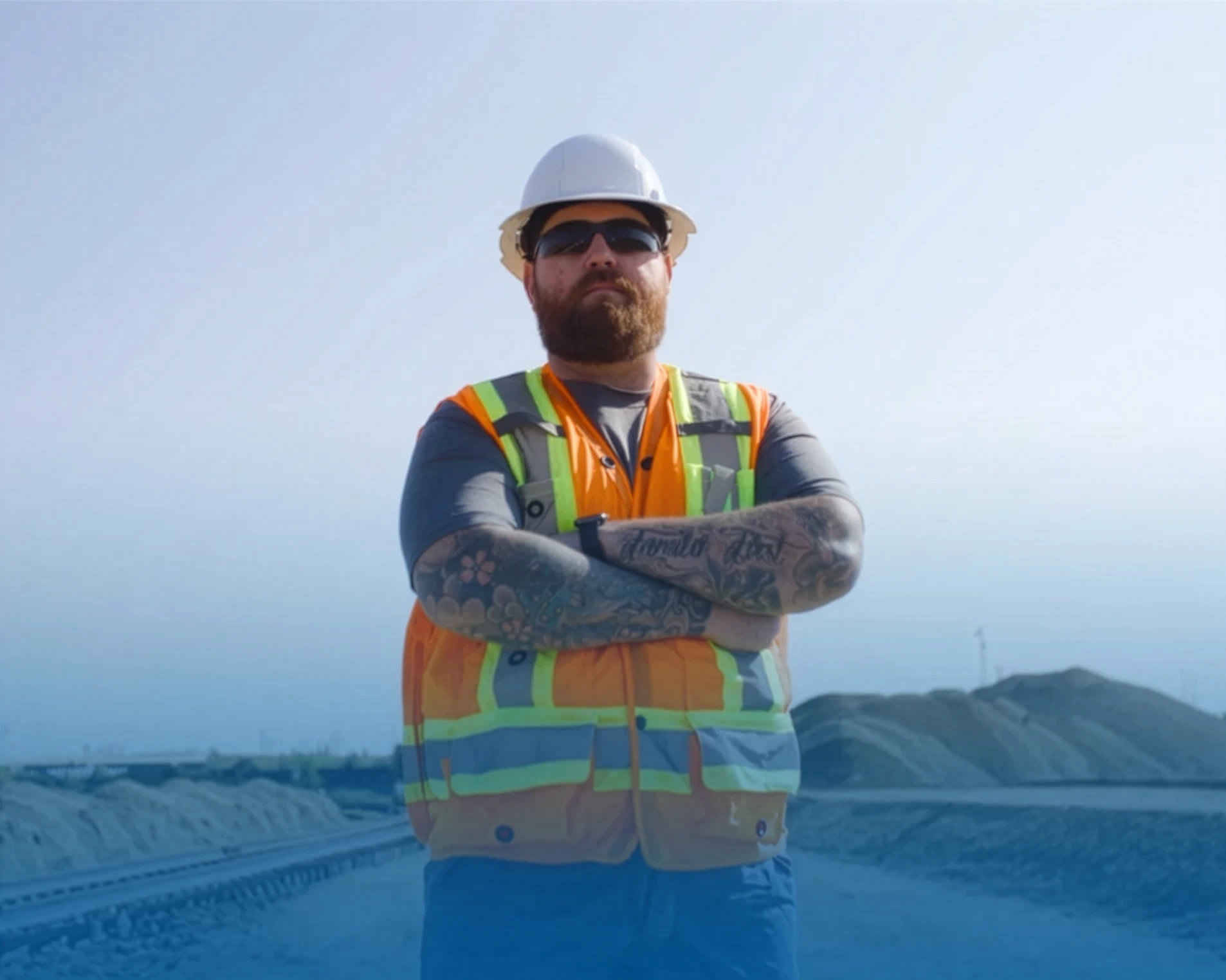 A bearded construction worker with tattoos, wearing a white hard hat, sunglasses, and a reflective orange safety vest, stands with arms crossed on a dirt worksite with mounds in the background.